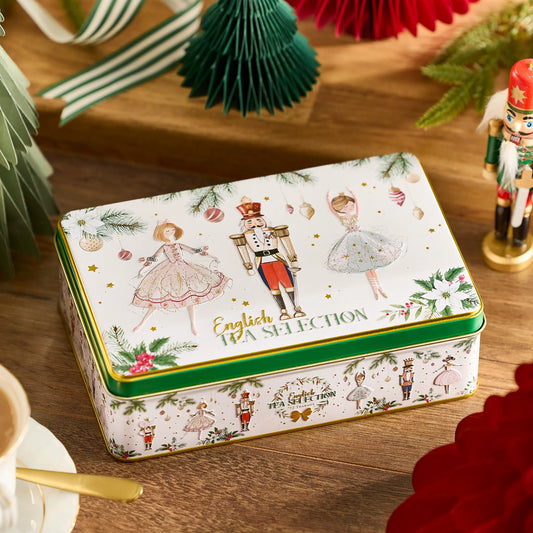 A decorative rectangular tin box labeled 'English Tea Selection' sits on a wooden table. The white tin has green trim and an illustrated scene with ballerinas, nutcrackers, holly, pinecones, and small tea-themed motifs.