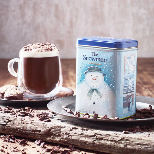 Tea canister with snowman design next to a cup of hot chocolate and cookies on a wooden surface.
