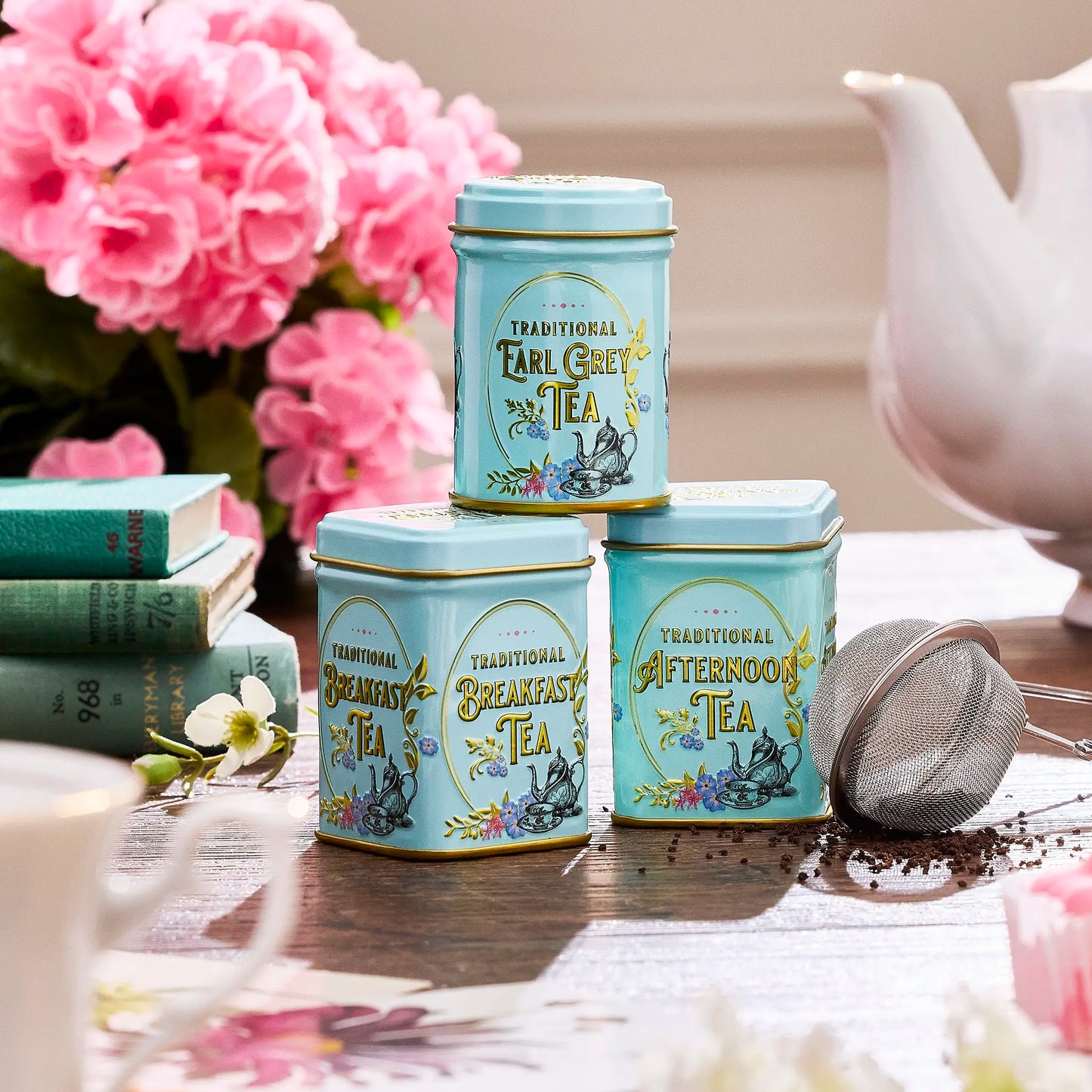 Three tea tins labeled 'Traditional Earl Grey Tea', 'Traditional Breakfast Tea', and 'Traditional Afternoon Tea' on a table with flowers and a teapot.