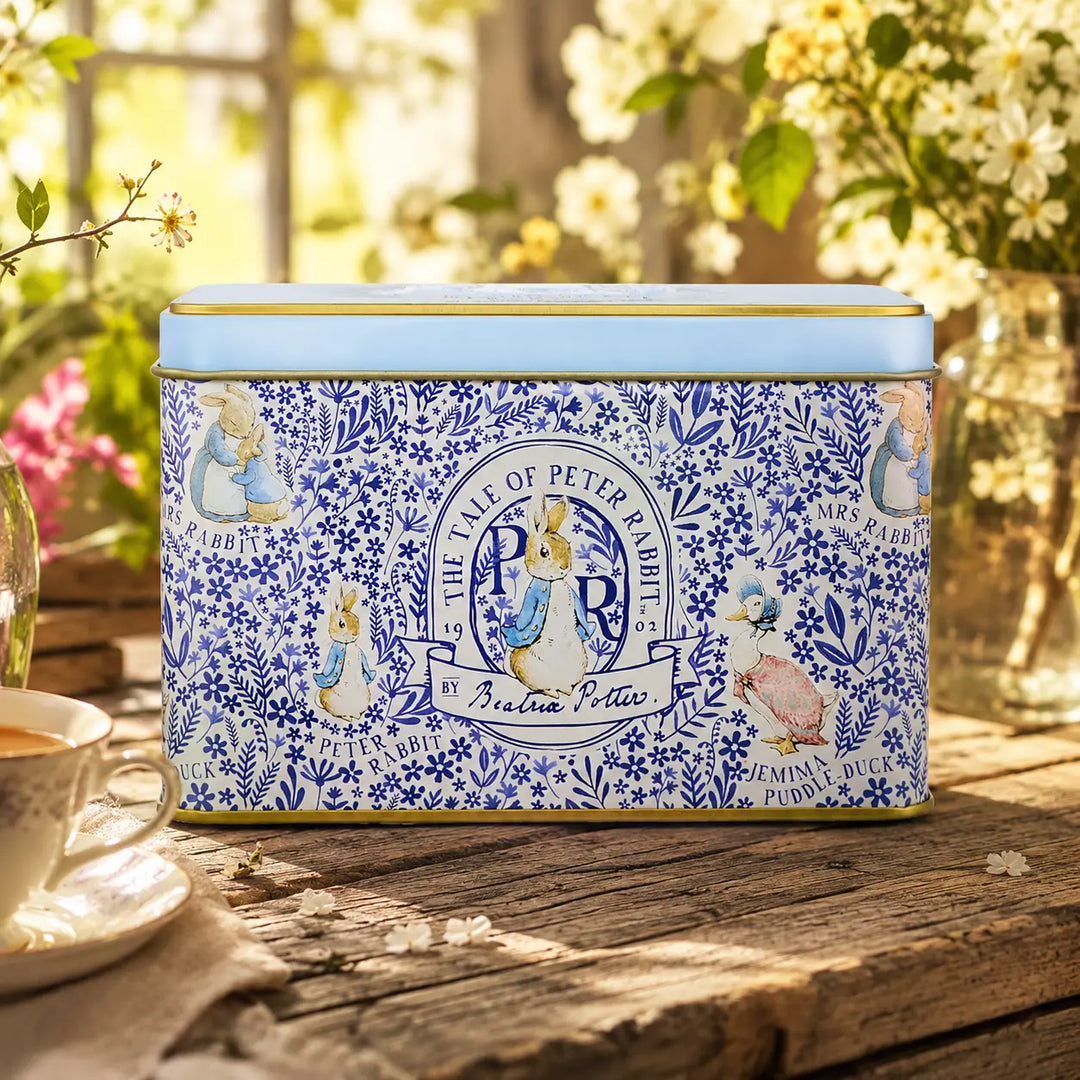 A rectangular blue and white decorative tin with Peter Rabbit illustrations sits on a rustic wooden table outdoors. The lid is light blue, and the front features blue floral patterns with Peter Rabbit, Jemima, and other characters.