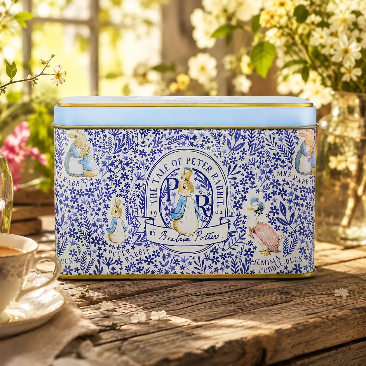 A rectangular blue and white decorative tin with Peter Rabbit illustrations sits on a rustic wooden table outdoors. The lid is light blue, and the front features blue floral patterns with Peter Rabbit, Jemima, and other characters.