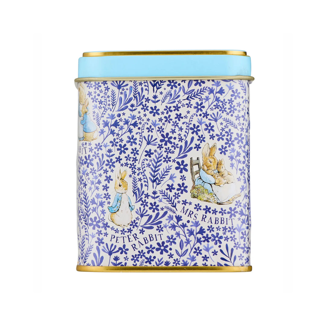 A cylindrical metal tin with a blue and white floral pattern featuring Peter Rabbit and Mrs. Rabbit illustrations, topped with a light blue lid and finished with gold-toned metal rims.