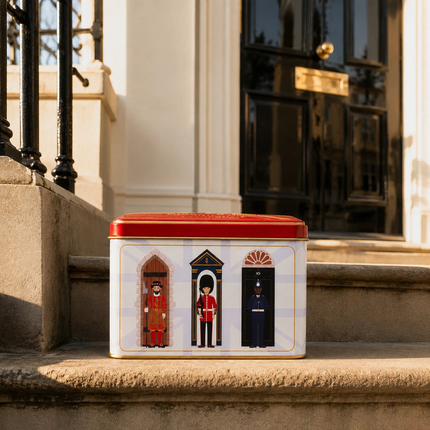 A rectangular tin with a red lid sits on stone steps in front of a building entrance. The tin has a white front panel showing three illustrated guard figures in red, gold, and dark blue uniforms.