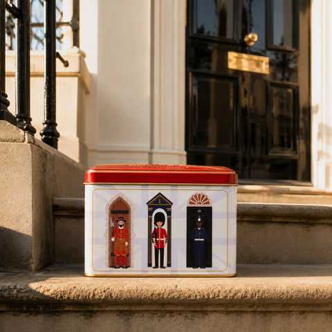A rectangular tin with a red lid sits on stone steps in front of a building entrance. The tin has a white front panel showing three illustrated guard figures in red, gold, and dark blue uniforms.