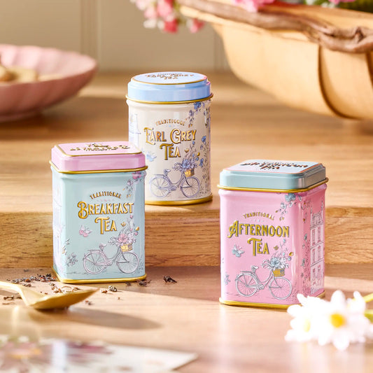 Three decorative metal tea tins arranged on a wooden surface in a softly styled tea-time setting. The tins are pastel-colored with floral bicycle illustrations and readable labels: Breakfast Tea in light blue and pink, Earl Grey Tea in cream and blue, and Afternoon Tea in pink and blue.