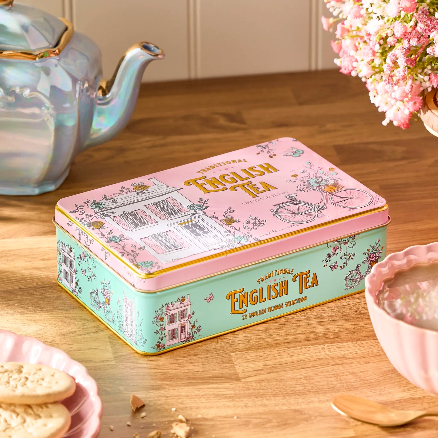 A decorative rectangular metal tin of traditional English tea sits on a wooden table, surrounded by a teapot, flowers, and a bowl of tea. The tin has a pink lid with gold lettering and a mint green side panel with floral illustrations.