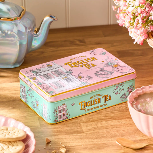 A decorative rectangular metal tin of traditional English tea sits on a wooden table, surrounded by a teapot, flowers, and a bowl of tea. The tin has a pink lid with gold lettering and a mint green side panel with floral illustrations.
