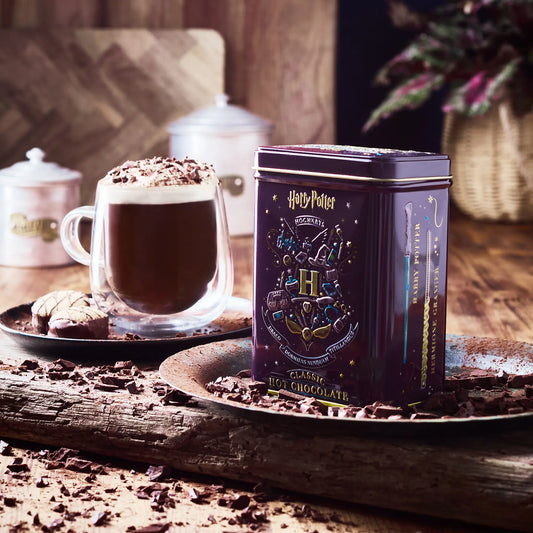 Hot chocolate with a Harry Potter-themed tin canister on a wooden surface with a cream topped hot chocolate drink, with chocolate shavings.
