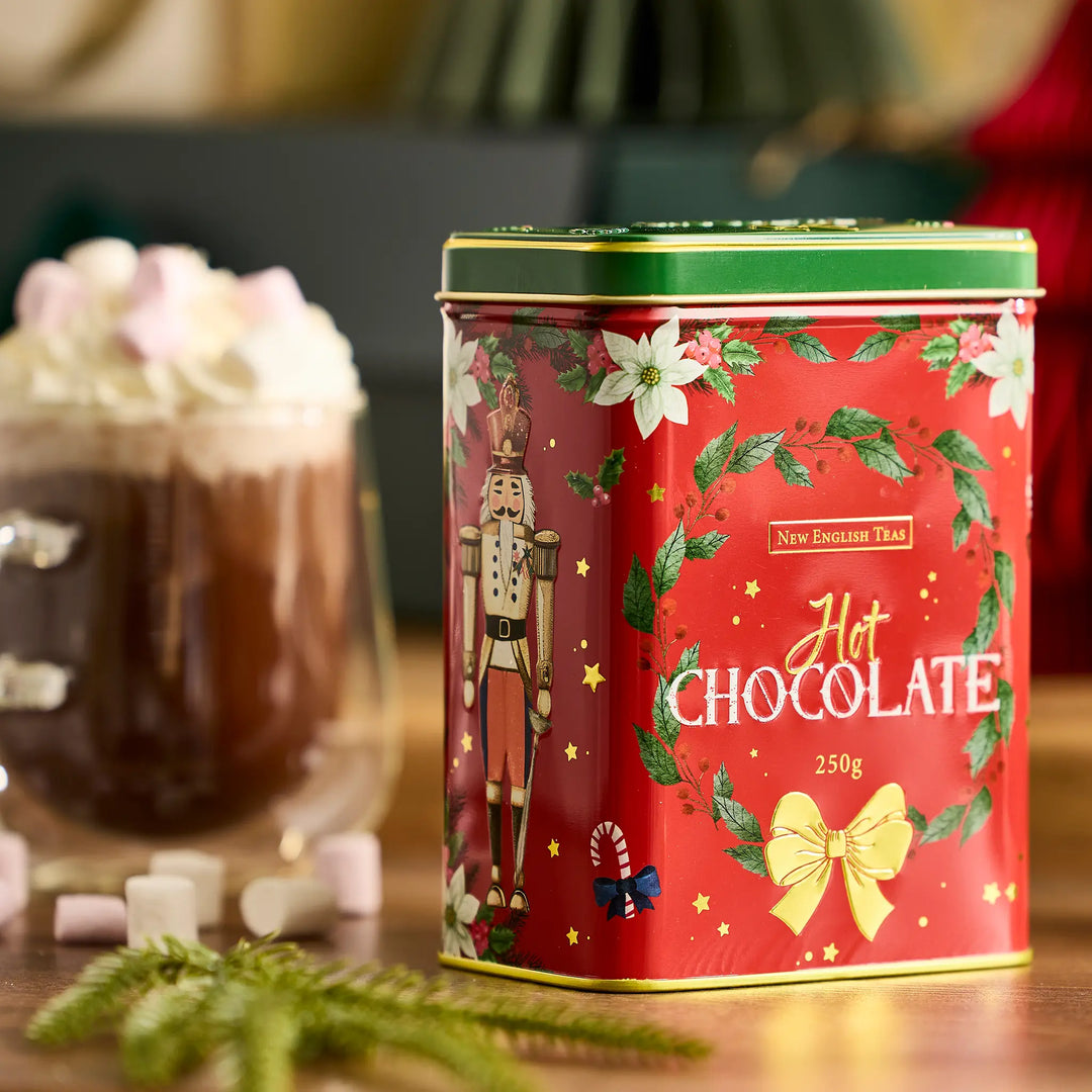 A red metal tin of New English Teas Traditional Hot Chocolate 250g, decorated with a nutcracker, white flowers, and green holly leaves, set on a wooden table next to a hot chocolate cup with whipped cream and marshmallows.