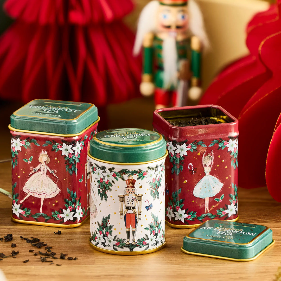 Three decorative Christmas-themed metal tea tins arranged on a wooden surface, with one tin open to show loose tea inside. The tins feature festive illustrations including ballerinas and a nutcracker, with red, white, and green color schemes and gold trim.