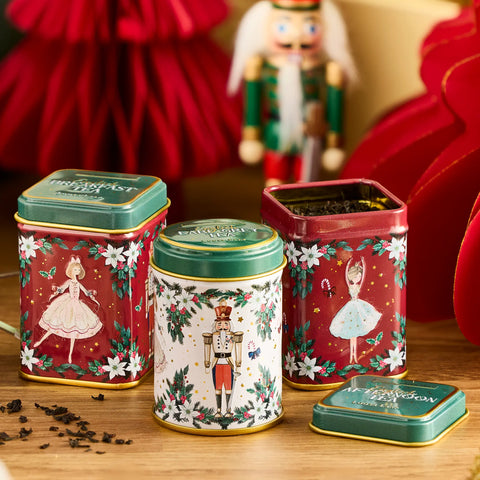 Three decorative Christmas-themed metal tea tins arranged on a wooden surface, with one tin open to show loose tea inside. The tins feature festive illustrations including ballerinas and a nutcracker, with red, white, and green color schemes and gold trim.