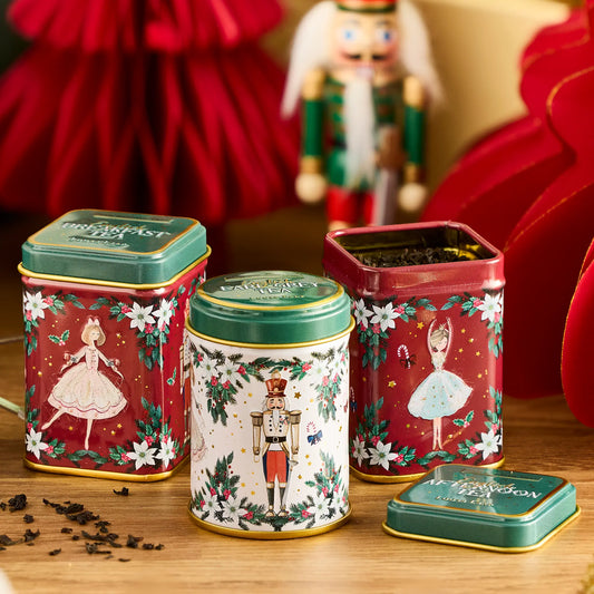 Three decorative Christmas-themed metal tea tins arranged on a wooden surface, with one tin open to show loose tea inside. The tins feature festive illustrations including ballerinas and a nutcracker, with red, white, and green color schemes and gold trim.