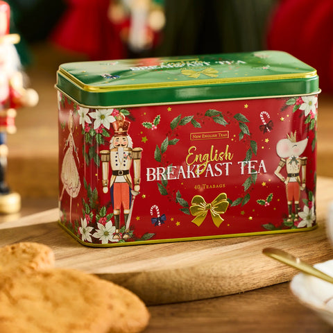 A red tin of New English Tea English Breakfast Tea sits on a wooden board in a festive setting. The tin features a green lid, a red body decorated with nutcracker illustrations, white flowers, green holly leaves, and gold accents.