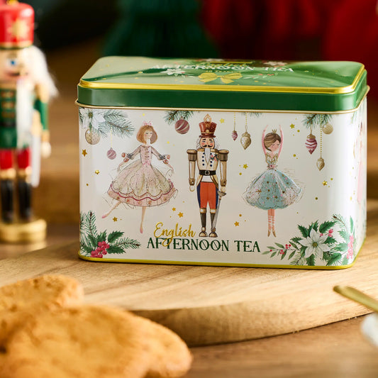 A rectangular metal tin of English Afternoon Tea with a green lid and a white front decorated with ballet dancers, nutcracker, ornaments, and floral illustrations. The tin sits on a wooden surface with cookies in the foreground and a blurred nutcracker figure behind.
