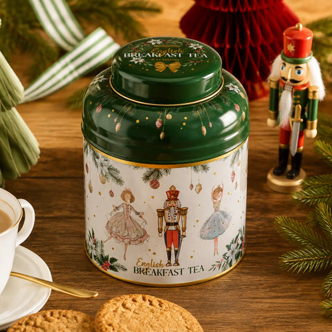 A decorative green and white tin of English Breakfast Tea sits on a wooden table surrounded by holiday decorations, including a cup and saucer, cookies, pine branches, and a nutcracker figurine.