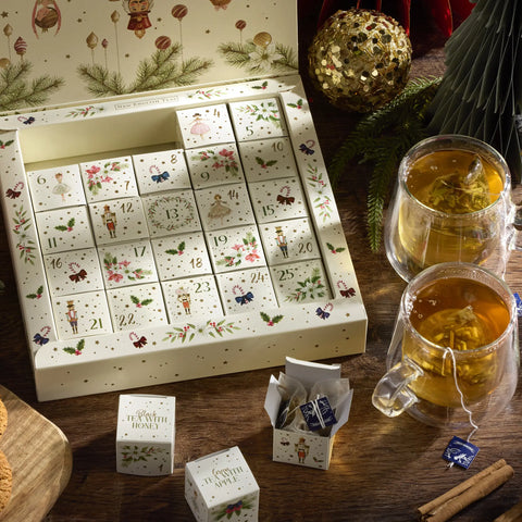 A Christmas advent tea gift set displayed on a wooden table, featuring an open box of illustrated individual tea bags, two glass mugs filled with brewed tea, and a few loose tea bags and cinnamon sticks around it.