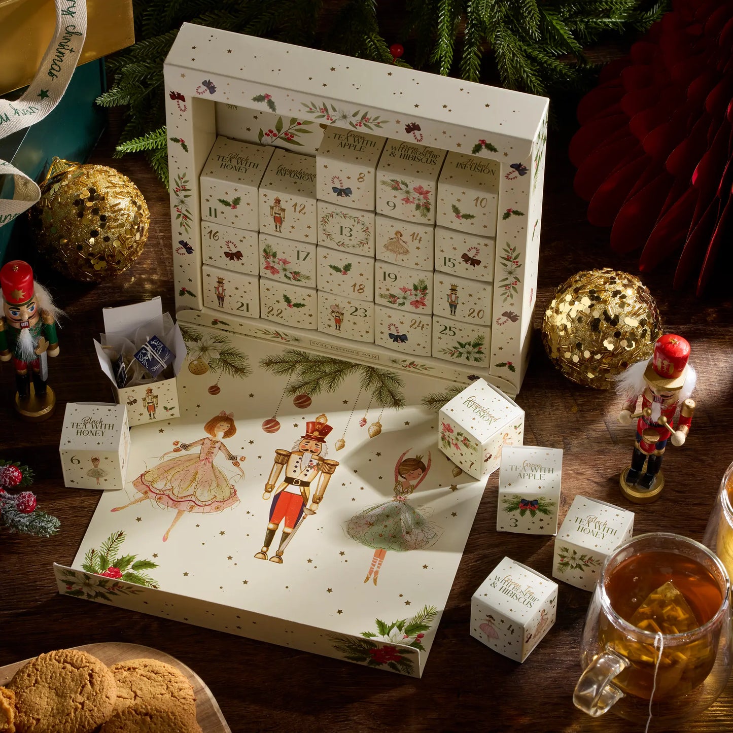 A festive Christmas advent calendar set displayed on a wooden table, featuring a large illustrated box with numbered compartments, several small matching boxes, nutcracker figurines, gold ornaments, cookies, and a glass mug of tea in a holiday setting.
