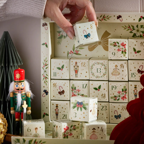 A hand opens one of a Christmas advent calendar cube boxes, revealing a small blue bow and gold ribbon illustration. The calendar features 25 numbered cream-colored boxes decorated with festive illustrations, set next to a nutcracker and a small green tree.