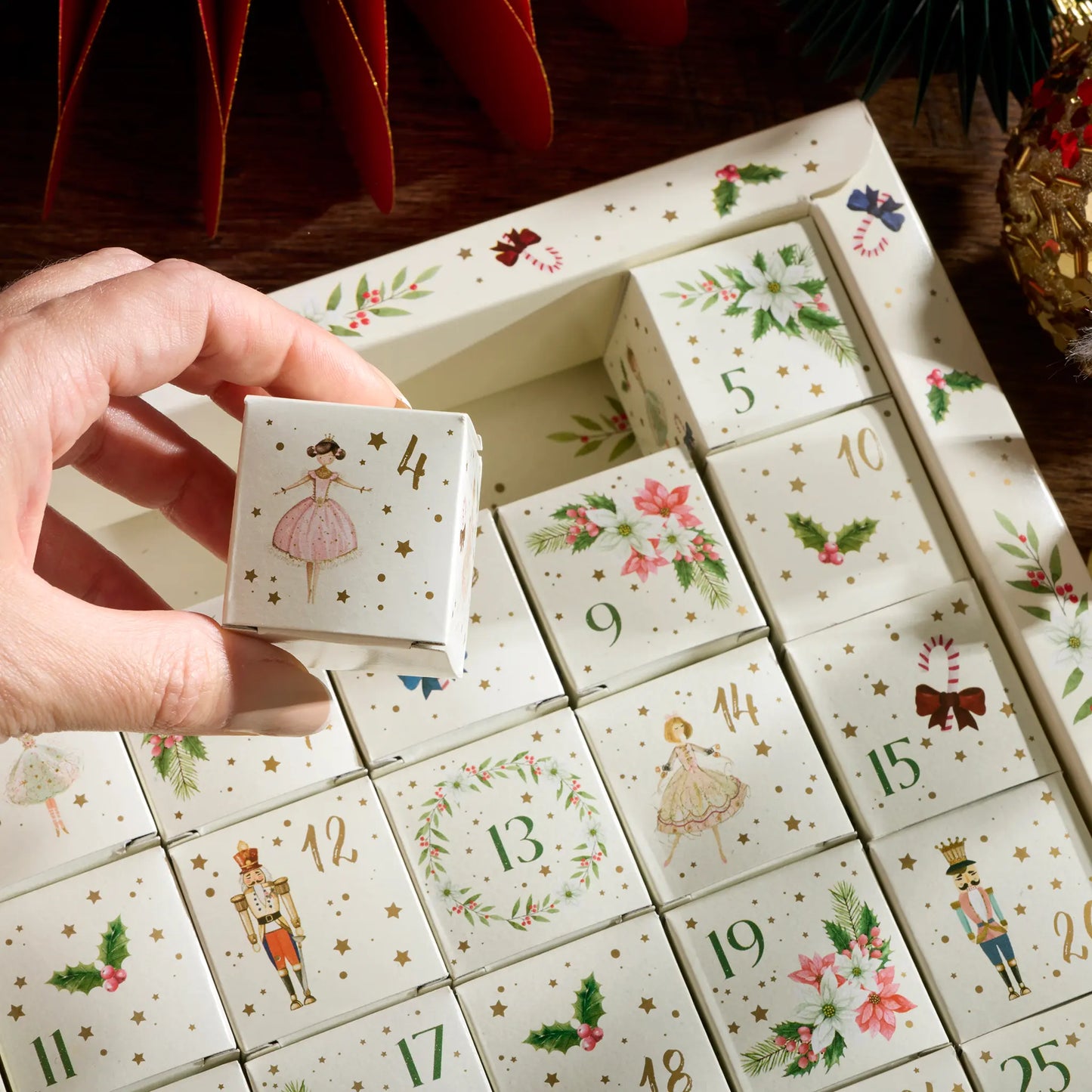 A hand holds a decorated white advent box cube numbered 4, with multiple matching advent cubes arranged in a tray below, each printed with a different holiday illustration and gold numbers.