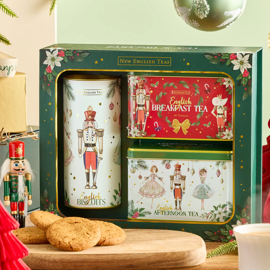 A boxed set of New English Tea holiday tea tins displayed in a gift box, with a large white nutcracker tin, a red Breakfast Tea tin, and a green Afternoon Tea tin. The set is styled on a wooden surface with English biscuits, a nutcracker figure, and holiday decorations.