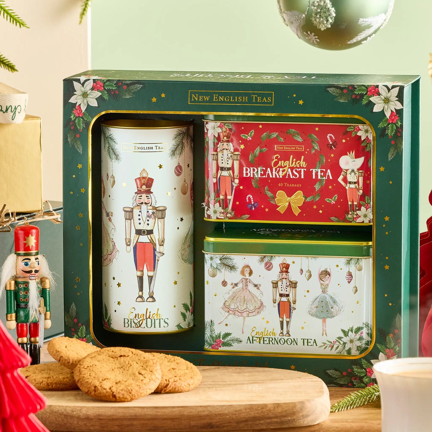 A boxed set of New English Tea holiday tea tins displayed in a gift box, with a large white nutcracker tin, a red Breakfast Tea tin, and a green Afternoon Tea tin. The set is styled on a wooden surface with English biscuits, a nutcracker figure, and holiday decorations.