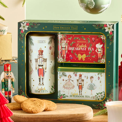 A boxed set of New English Tea holiday tea tins displayed in a gift box, with a large white nutcracker tin, a red Breakfast Tea tin, and a green Afternoon Tea tin. The set is styled on a wooden surface with English biscuits, a nutcracker figure, and holiday decorations.