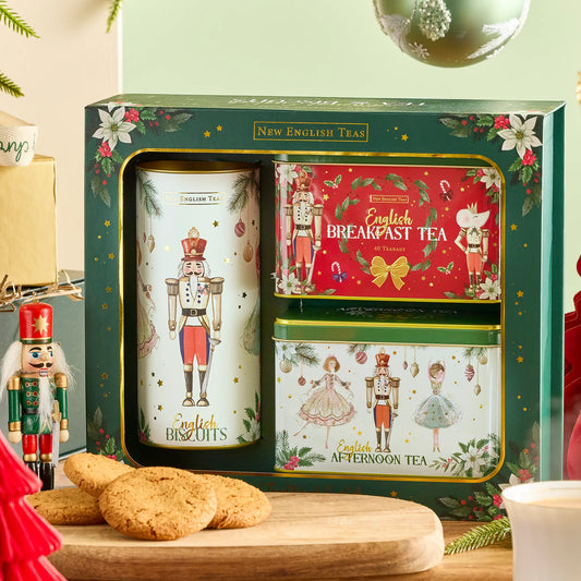 A boxed set of New English Tea holiday tea tins displayed in a gift box, with a large white nutcracker tin, a red Breakfast Tea tin, and a green Afternoon Tea tin. The set is styled on a wooden surface with English biscuits, a nutcracker figure, and holiday decorations.