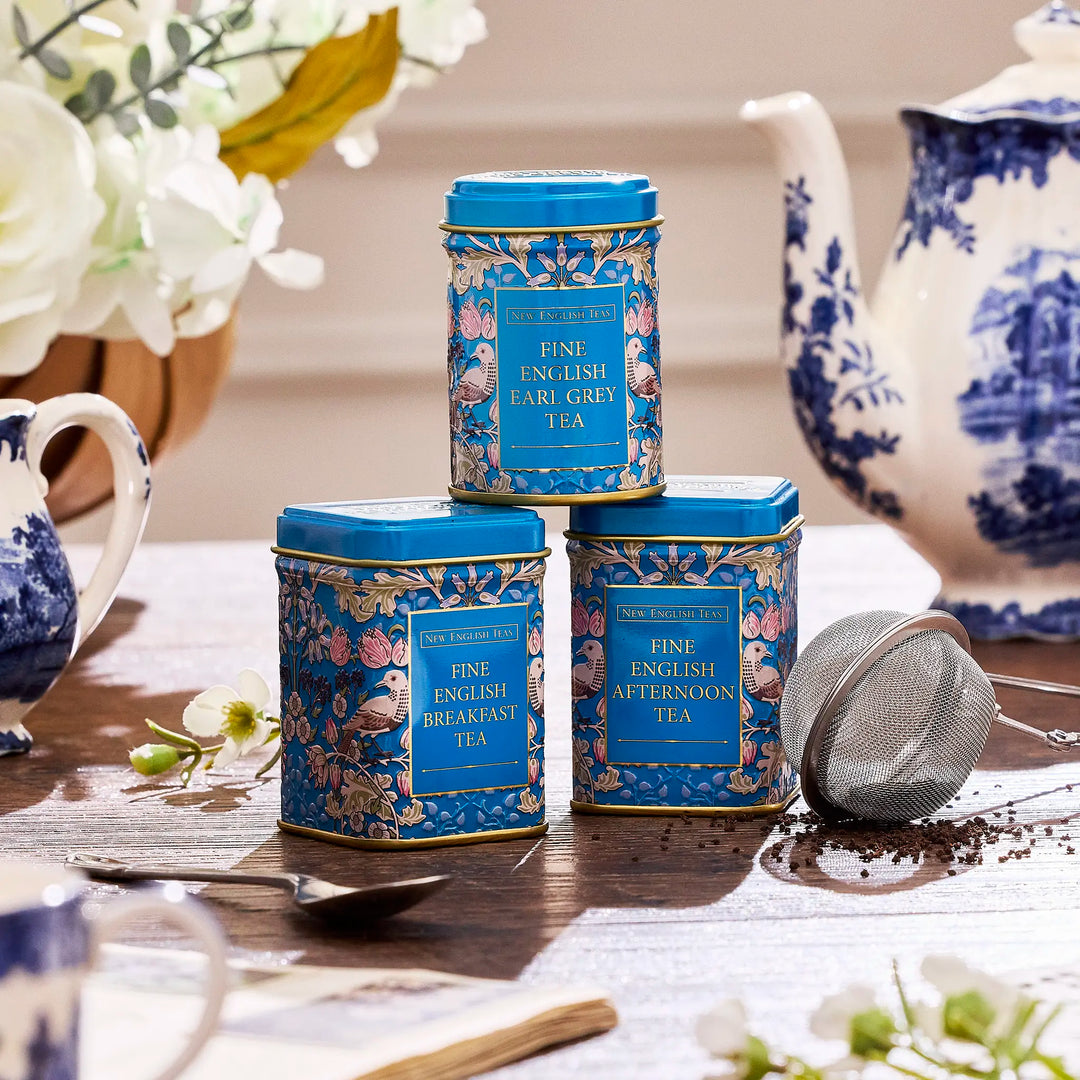 Three blue tea tins with floral designs on a table with teacup and teapot.