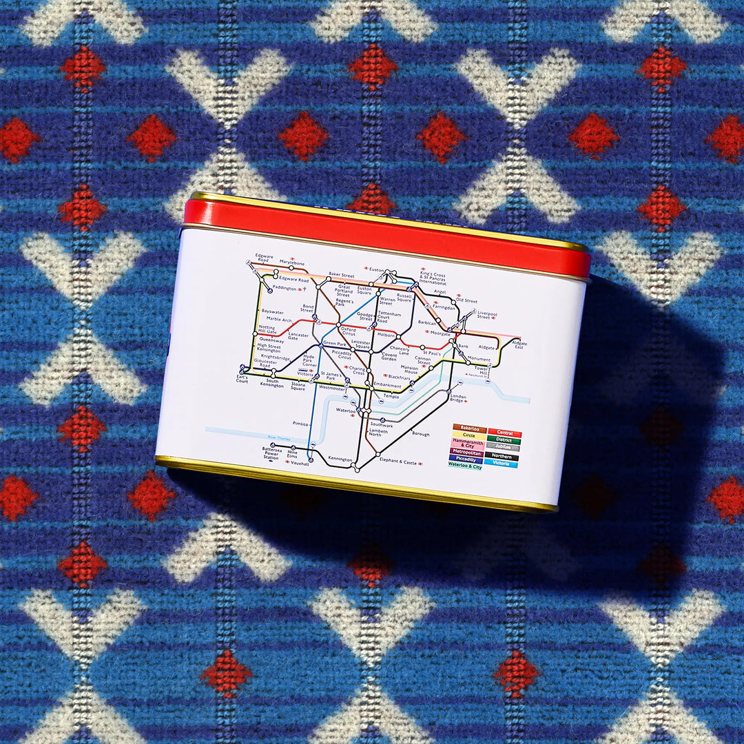 A rectangular tin with a white label printed with a colorful London tube map, featuring a red top rim and gold bottom rim, placed on a blue patterned knit Transport for London Moquette design.