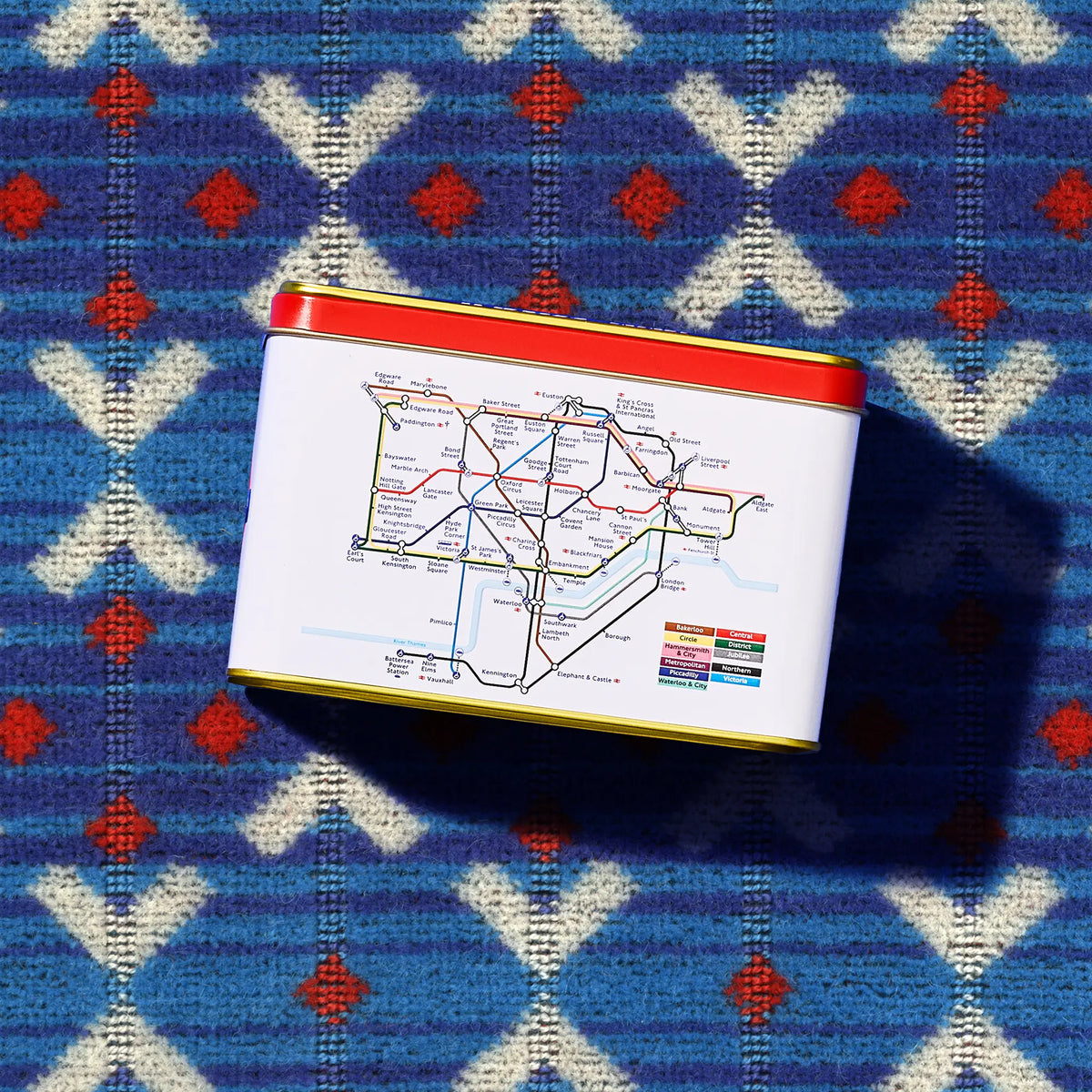 A rectangular tin with a white label printed with a colorful London tube map, featuring a red top rim and gold bottom rim, placed on a blue patterned knit Transport for London Moquette design.