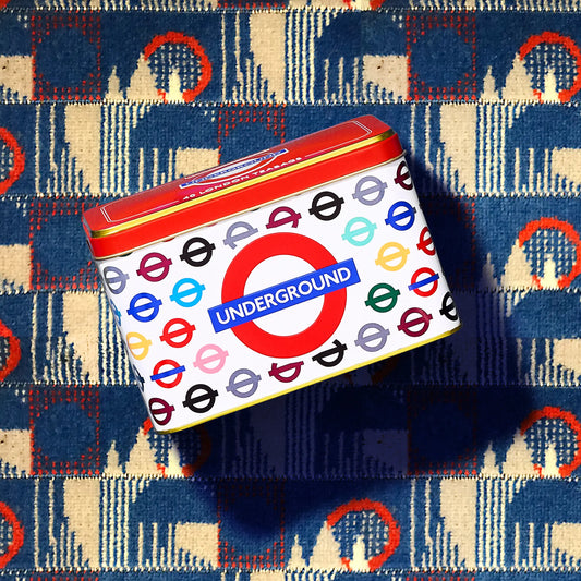A square metal tin with a London Underground roundel and tube map design, placed on top of a blue and cream patterned textile background.