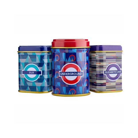 Three decorative metal tins with London Underground-themed designs on a plain white background. The left tin is blue with geometric patterns and the DLR logo, the center tin is blue with a red lid and the Underground roundel, and the right tin is gray with striped patterns and the Elizabeth Line logo.