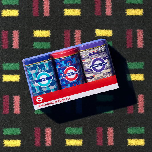 A boxed set of three London-themed tea tins arranged diagonally on a dark textured surface with colorful striped patterns. The tins feature London Underground-style logos and colors, including blue, red, and purple, with labels reading DLR, Underground, and Elizabeth Line.