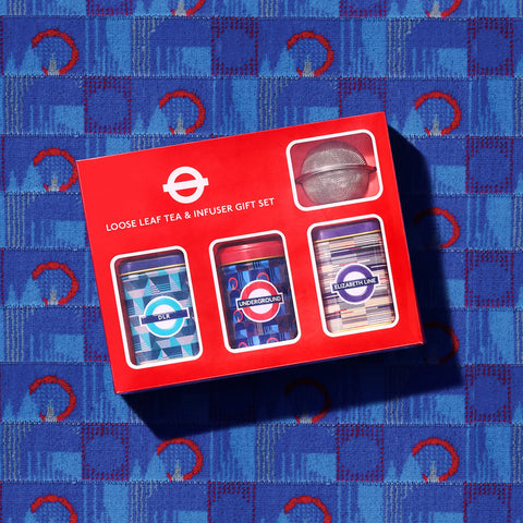 A red gift box labeled 'Loose Leaf Tea & Infuser Gift Set' with three tea infusers visible through cutouts, placed on a blue patterned fabric background.